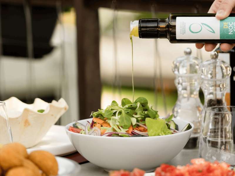 Eine Hand gießt natives Olivenöl extra vom Gardasee aus einer Flasche über einen Teller Salat
